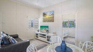 A living room with a white sofa, a TV on a white stand, a painting of a beach scene, and a view through a window. - Evans Above - 1-5 Wattle Street (EVANS HEAD)