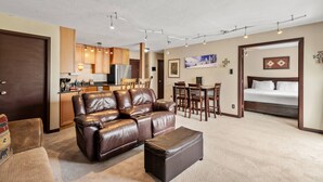 Una sala de estar moderna con un sofá de cuero marrón, un reposapiés a juego, un comedor con sillas de madera y una cocina al fondo.