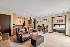 A modern living room with a brown leather sofa, a matching ottoman, a dining area with wooden chairs, and a kitchen in the background.