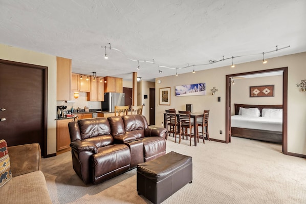 A modern living room with a brown leather sofa, a matching ottoman, a dining area with wooden chairs, and a kitchen in the background.