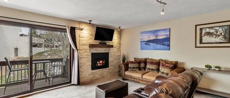 A living room with a fireplace, a sofa, a television, and a large window overlooking a deck.
