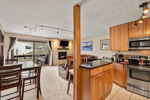 A modern kitchen with wooden cabinets, a built-in oven, and a microwave. There is a dining area with a table and chairs, and a fireplace in the background.