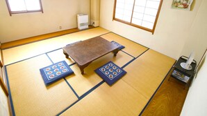 Une pièce avec un sol recouvert de tatamis, une table en tatami, deux tatamis aux motifs traditionnels, un radiateur et une petite étagère avec quelques objets.