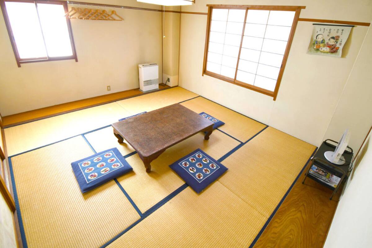 Une pièce avec un sol recouvert de tatamis, une table en tatami, deux tatamis aux motifs traditionnels, un radiateur et une petite étagère avec quelques objets.