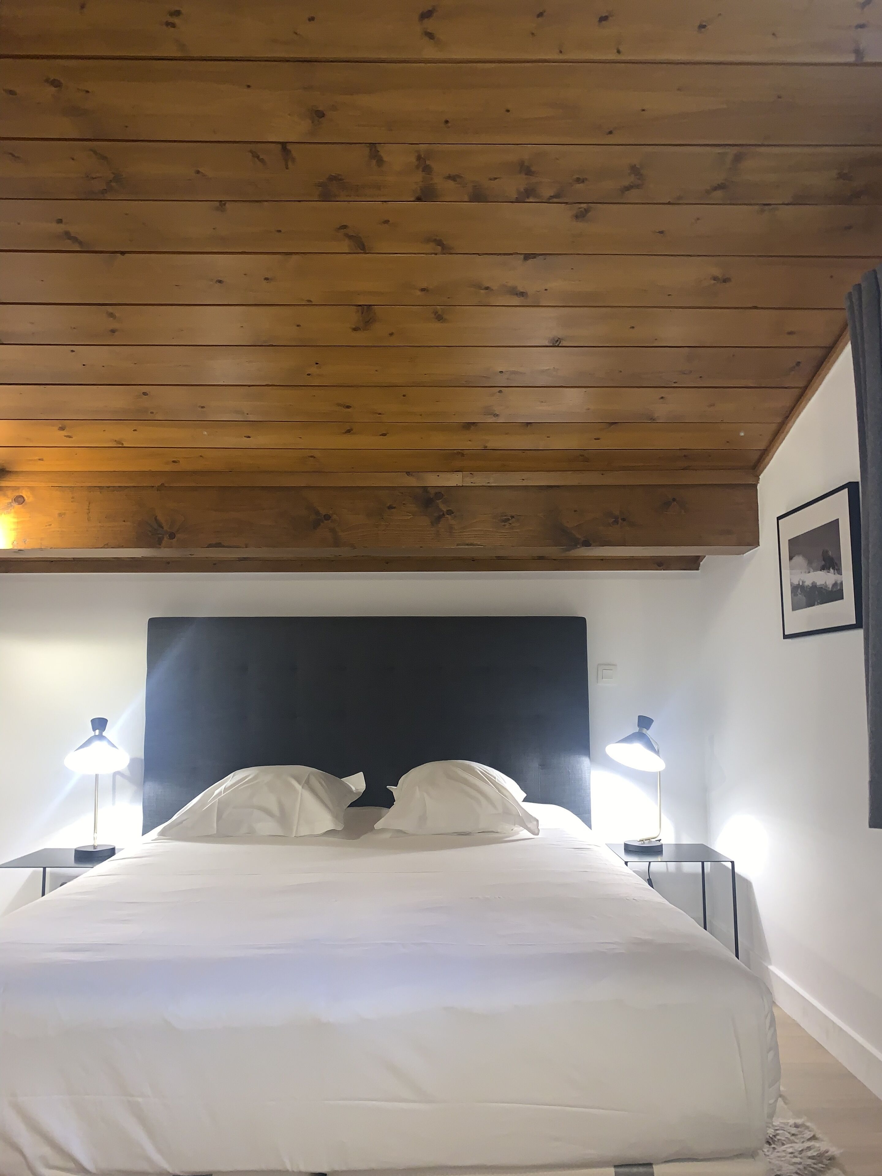 A bedroom with a wooden ceiling, a black headboard, white bedding, and two bedside lamps.