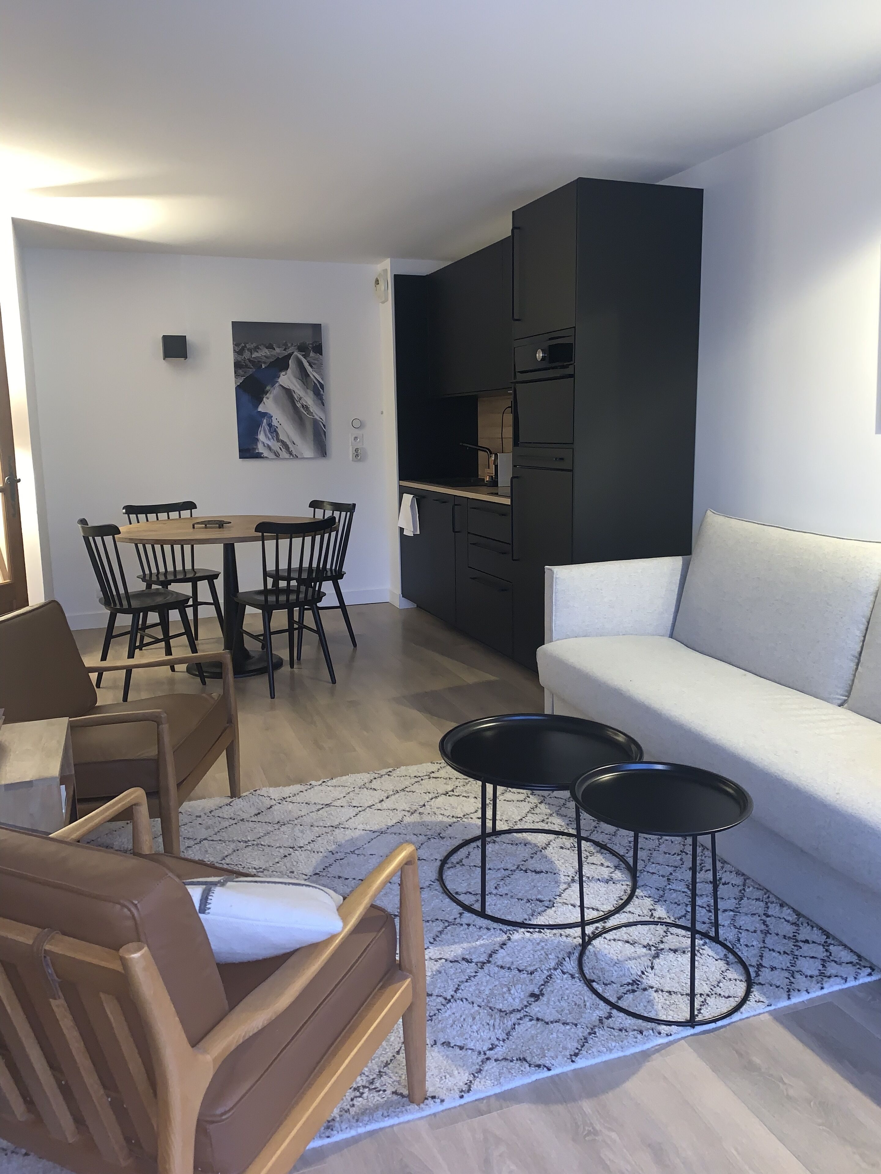 Una sala de estar moderna con un sofá, dos mesitas auxiliares negras, una silla de madera, una mesa de comedor con sillas y una cocina con armarios y electrodomésticos negros.