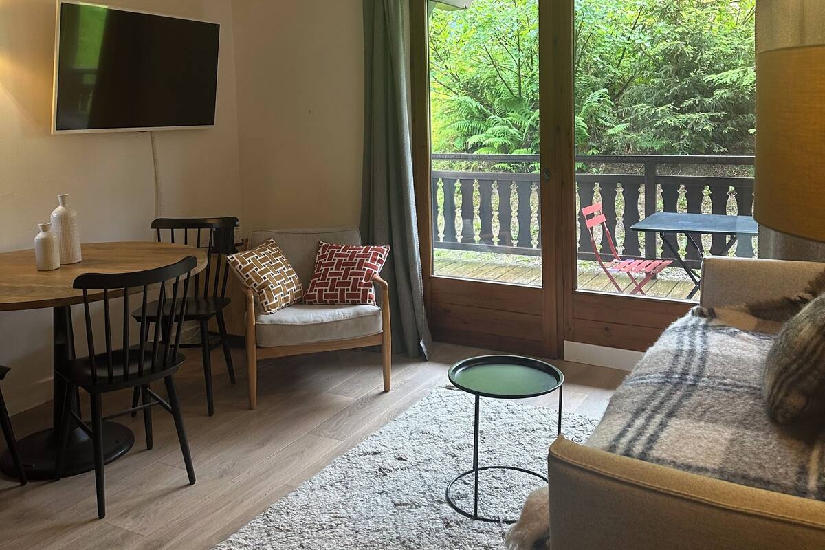 Uma sala de estar com televisão, uma mesa de jantar com cadeiras, um sofá e vista para uma varanda com mobiliário externo.