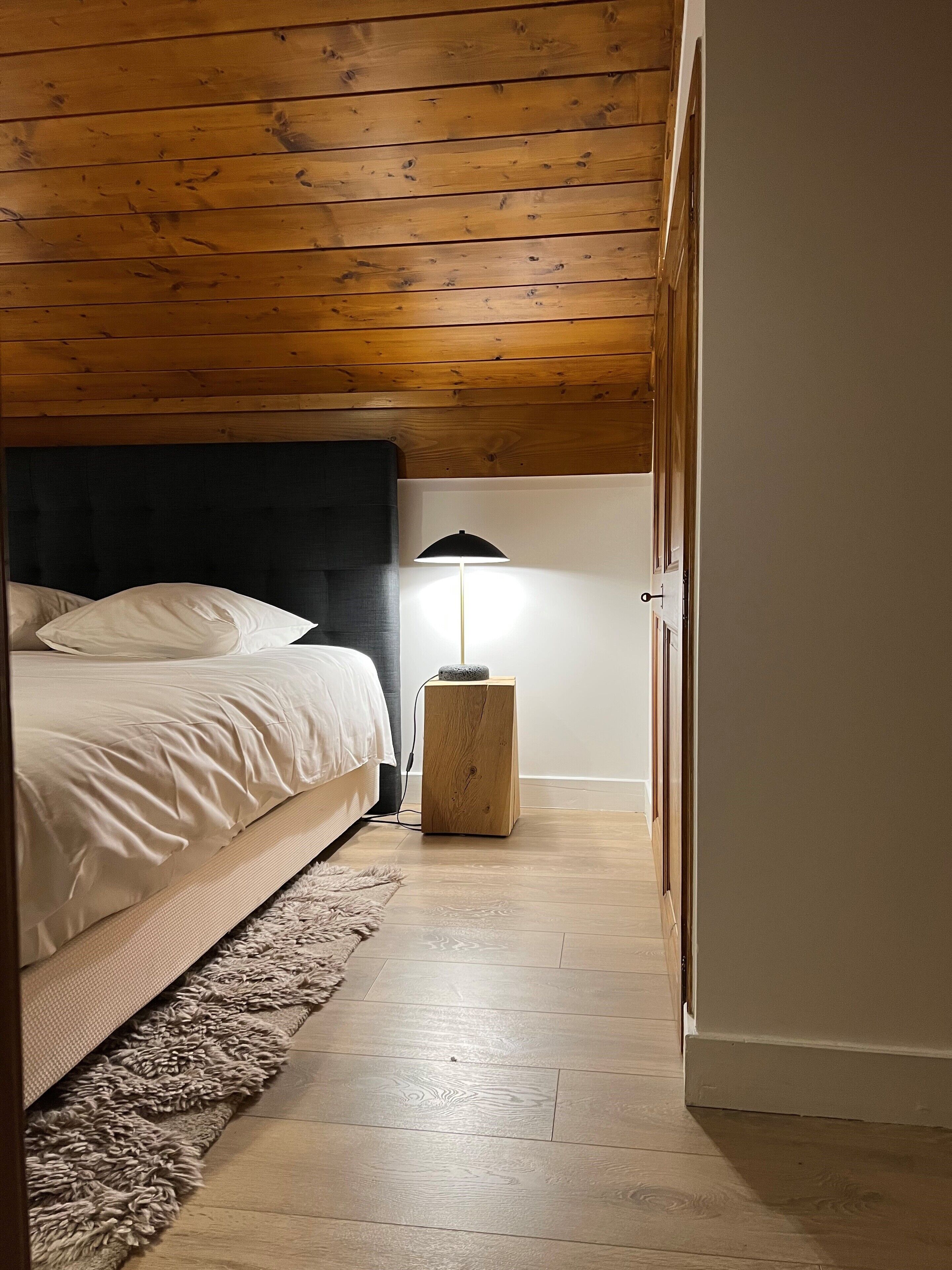 Ein Schlafzimmer mit Holzbalkendecke, einem Bett mit weißer Bettwäsche, einem kleinen Nachttisch mit Lampe und einem flauschigen Teppich auf dem Boden.