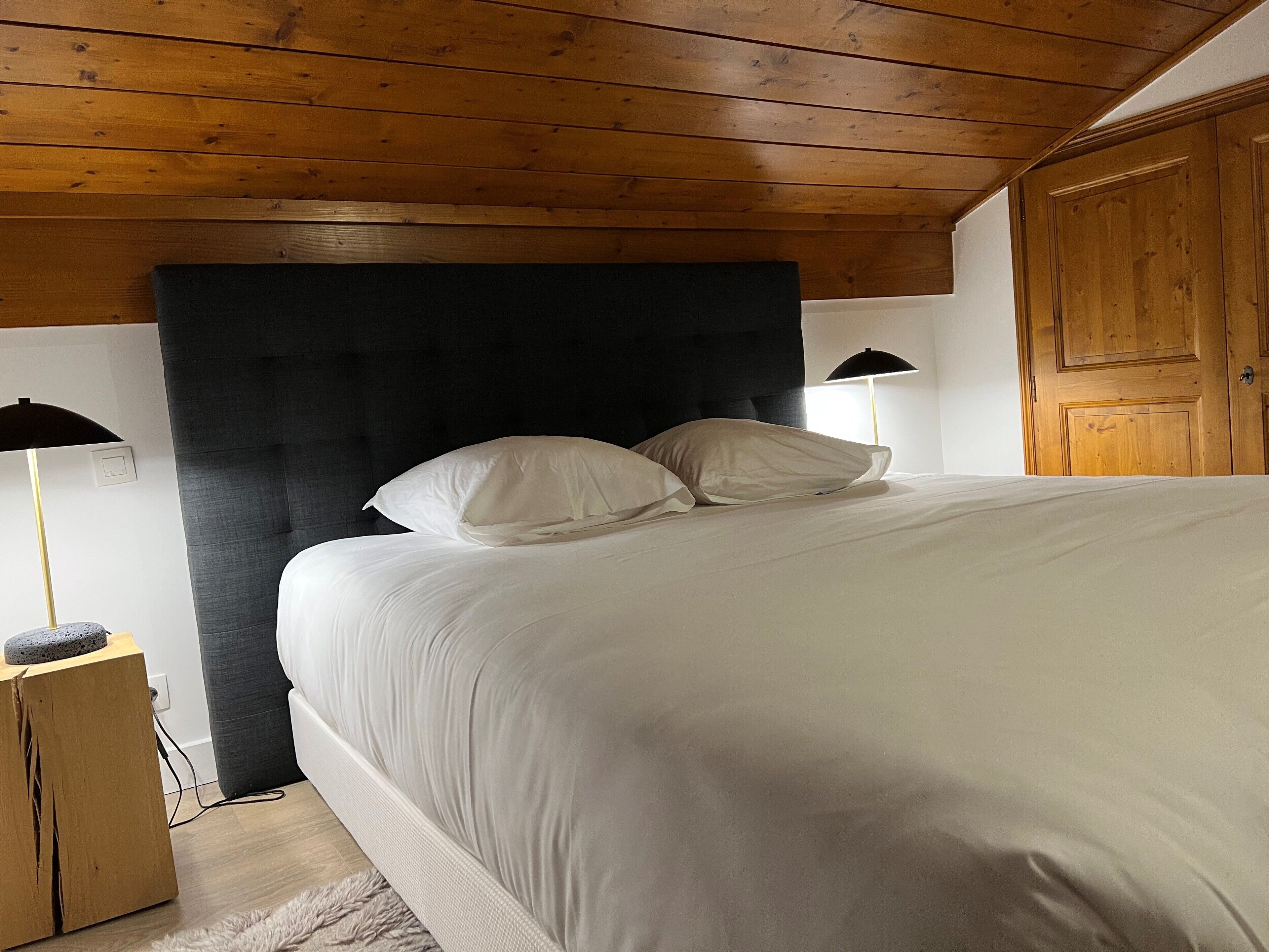 A bedroom with a wooden ceiling, a large bed with white bedding, two bedside lamps, and a wooden wardrobe.
