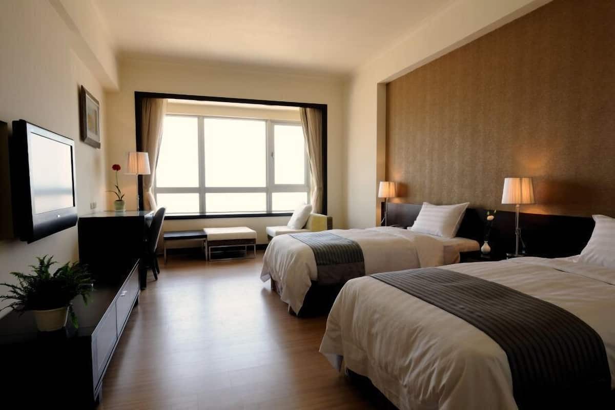 Habitación de hotel con dos camas, un escritorio, una silla, un televisor y un ventanal con cortinas.