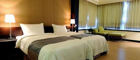 Habitación de hotel con dos camas grandes, una zona de estar con una silla, una lámpara y un ventanal con cortinas.
