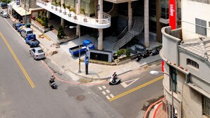 Ein mehrstöckiges Hotel im modernen Design mit Balkonen auf jeder Etage, einem zentralen Eingang mit Treppen und einem Parkplatz für Autos und Roller.