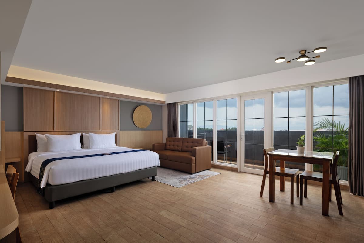 A modern hotel room with a large bed, a sofa, a dining table, and a balcony with a view.