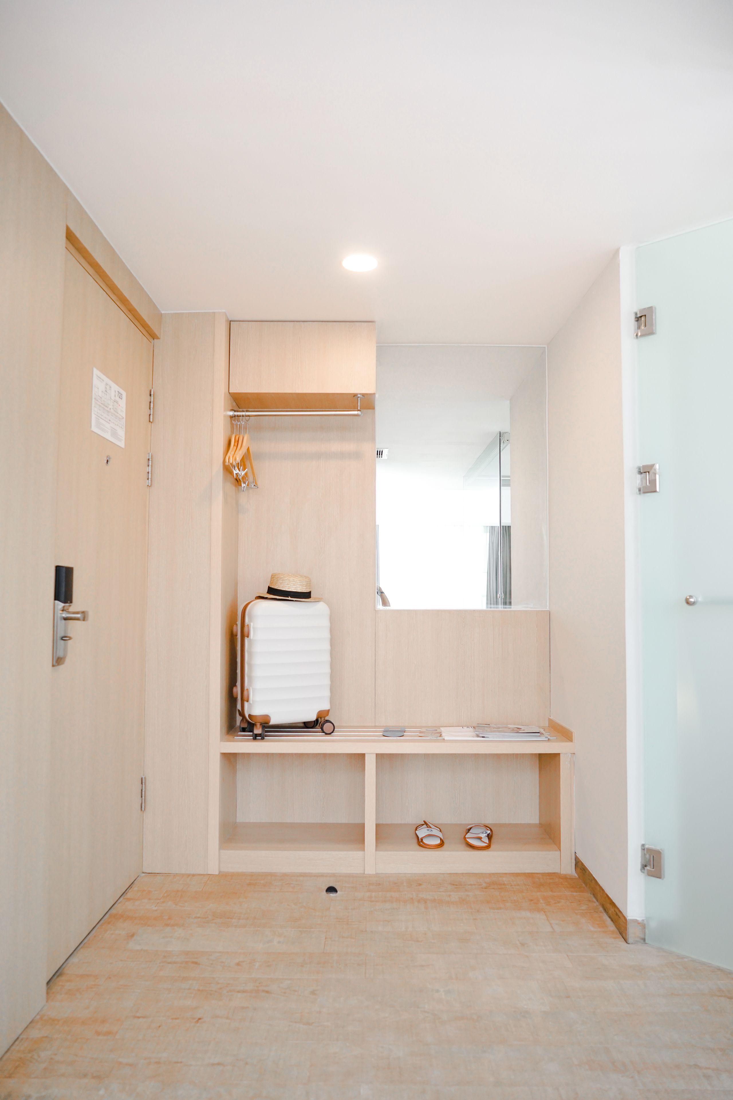 A hotel room with a wooden floor, a white suitcase, a hat, and a pair of sandals on a shelf.