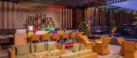 Un área de lounge exterior bien iluminada con mobiliario de mimbre, un bar con estantes llenos de botellas y una pared decorativa.