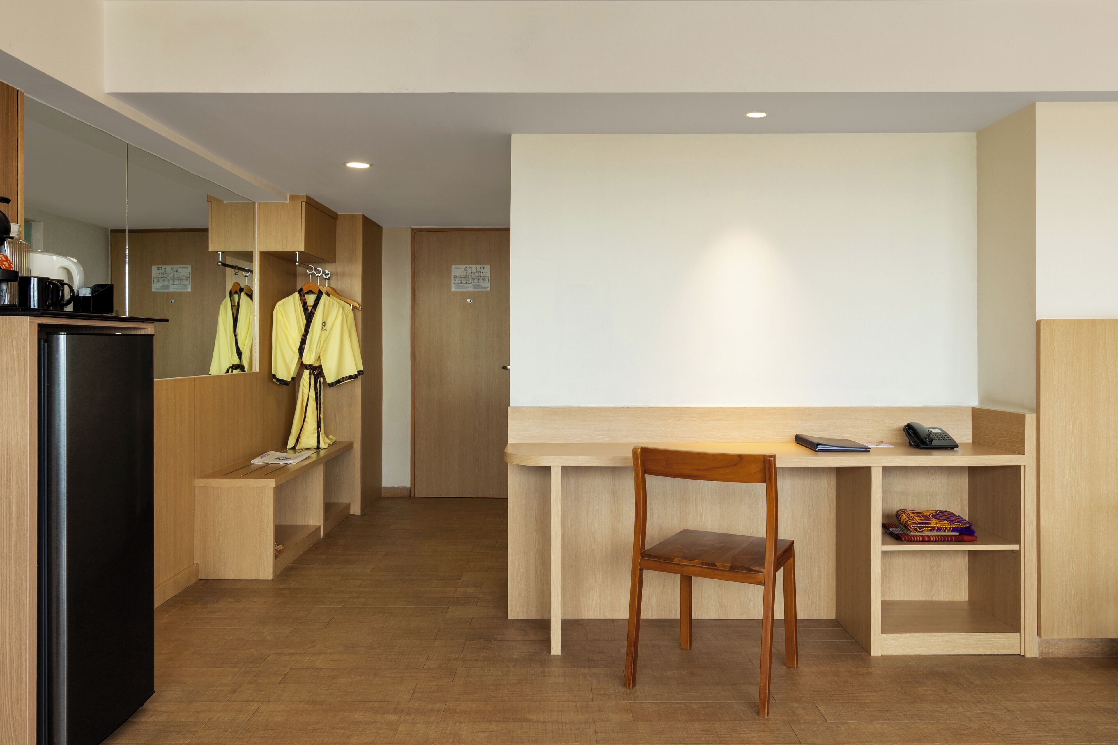 Une chambre d’hôtel avec un bureau en bois, une chaise, une petite étagère avec un carnet, et un porte-manteau avec deux vestes.