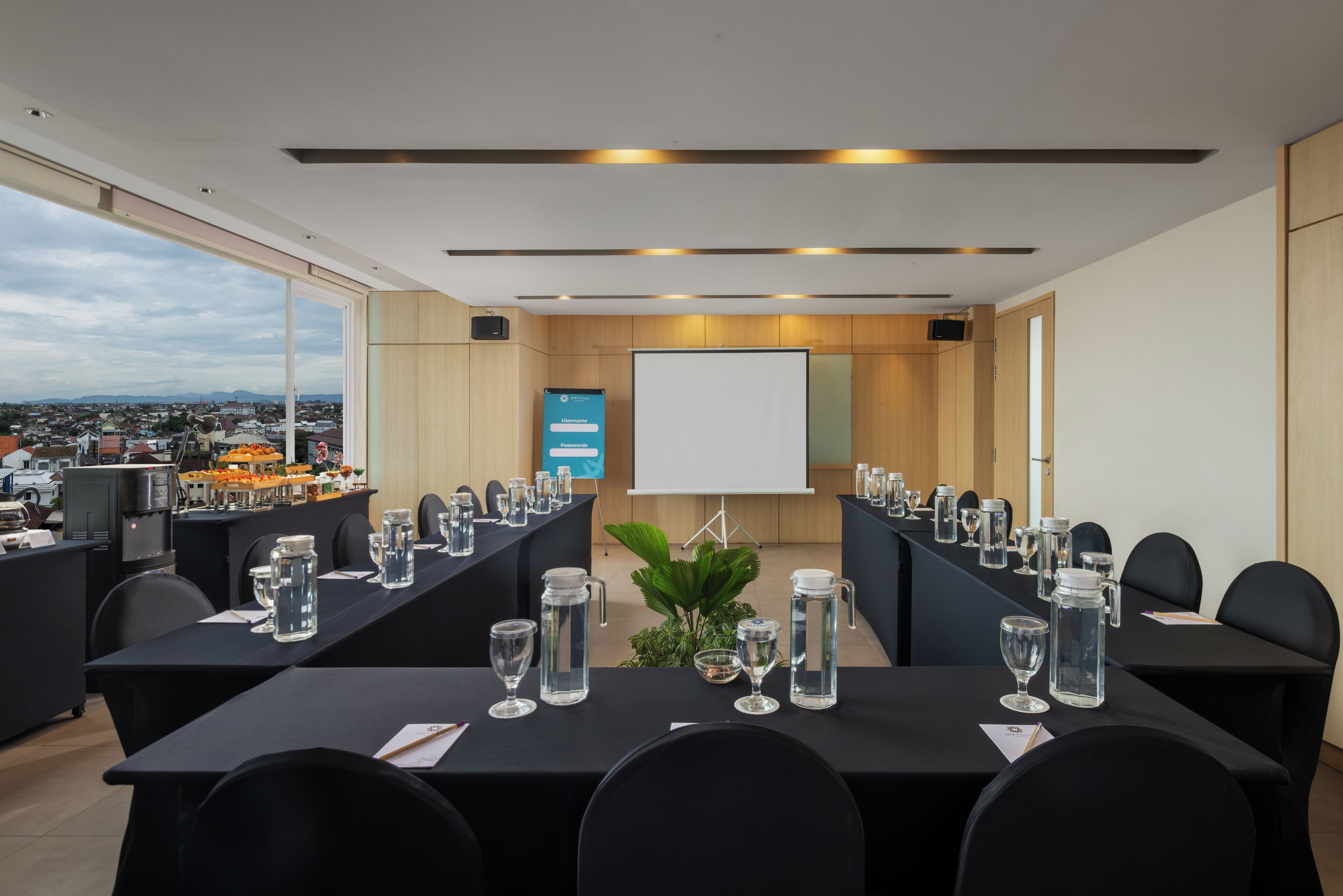 A conference room with a large table, chairs, a projector screen, and a view of a cityscape through large windows.