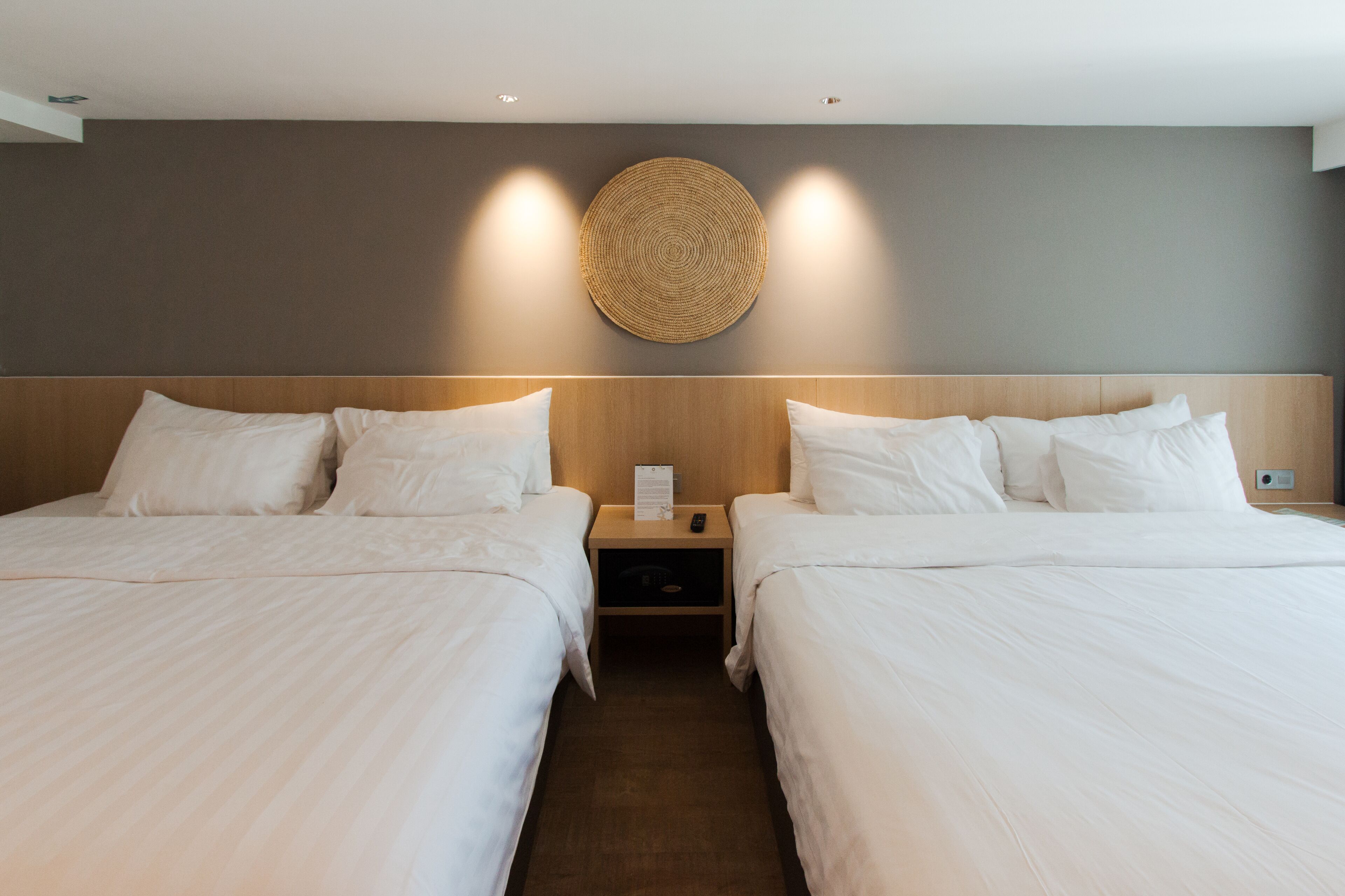 A hotel room with two beds, a wooden headboard, a woven wall decoration, and a nightstand with a remote control.