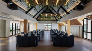 Een vergaderzaal met lange tafels, zwarte stoelen en een hoog plafond met houten balken en hangplanten.