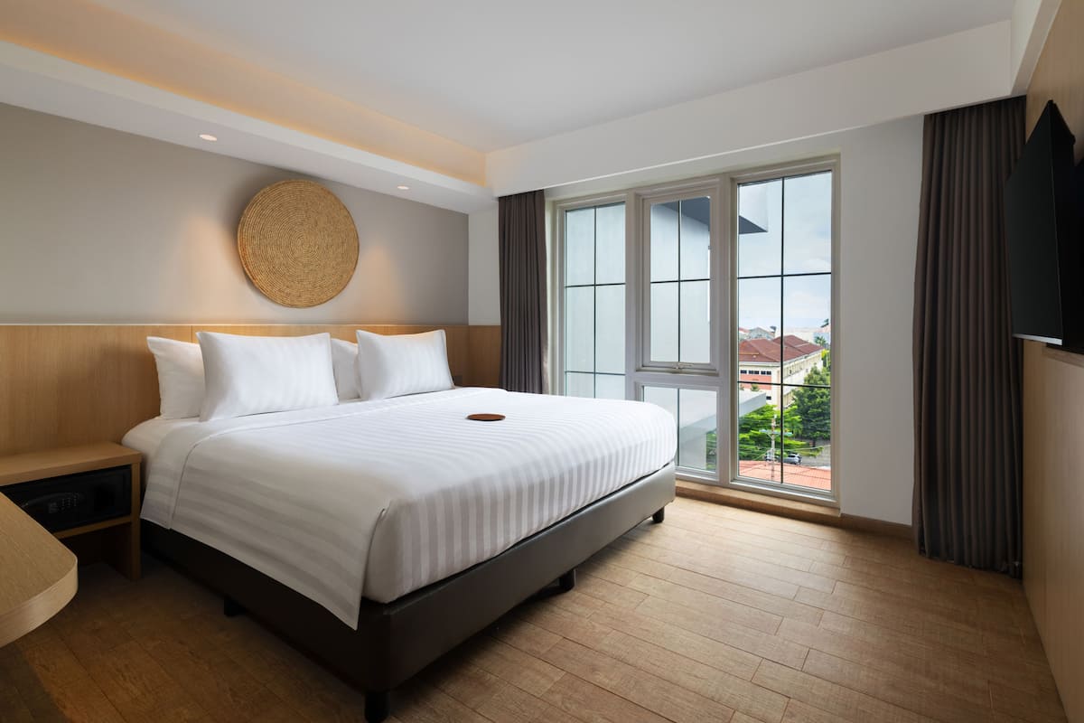 A hotel room with a large bed, a bedside table, a flat-screen TV, and a view of the city through the window.