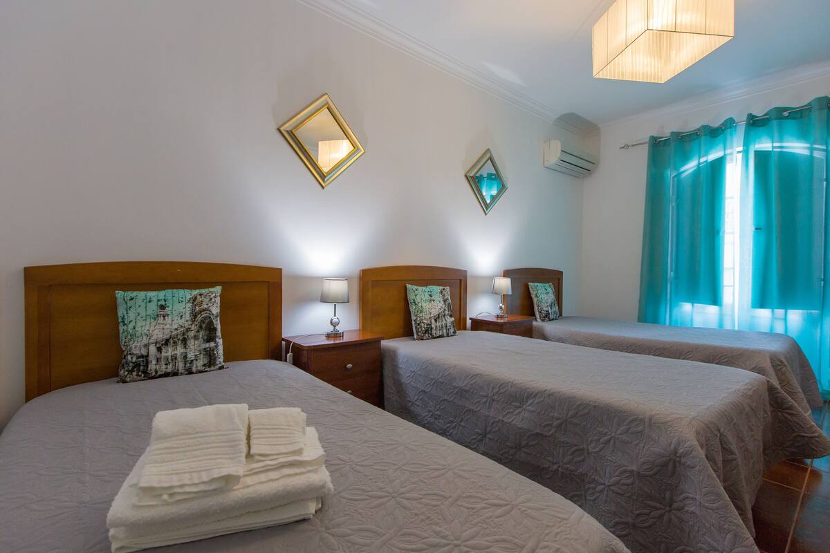 A hotel room with two beds, each with a wooden headboard, a small bedside table with a lamp, and a wall-mounted air conditioner.