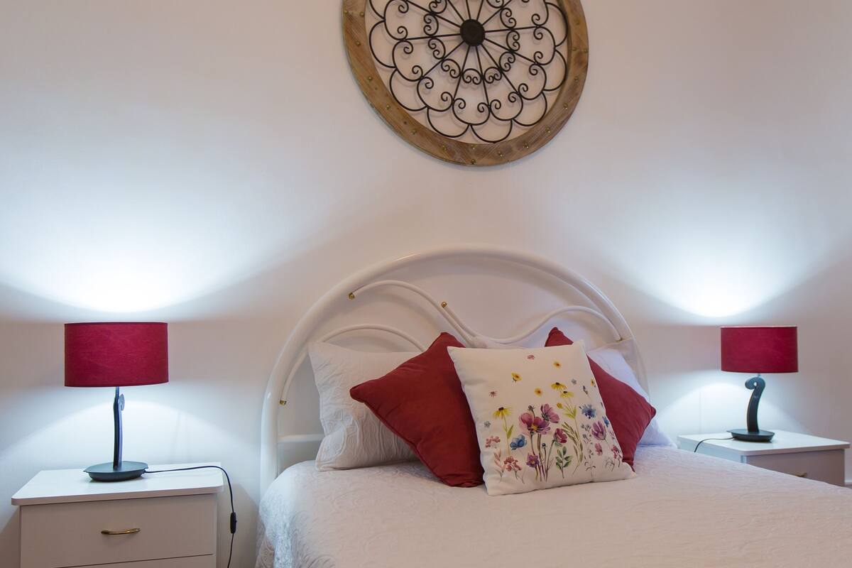 A bedroom with a white bed, red and floral pillows, a white nightstand with a lamp, and a decorative wall piece.