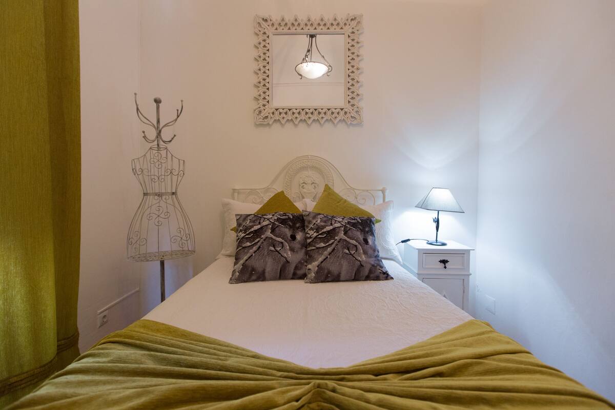 A neatly made bed with a green blanket, a bedside lamp, and a decorative mirror on the wall.