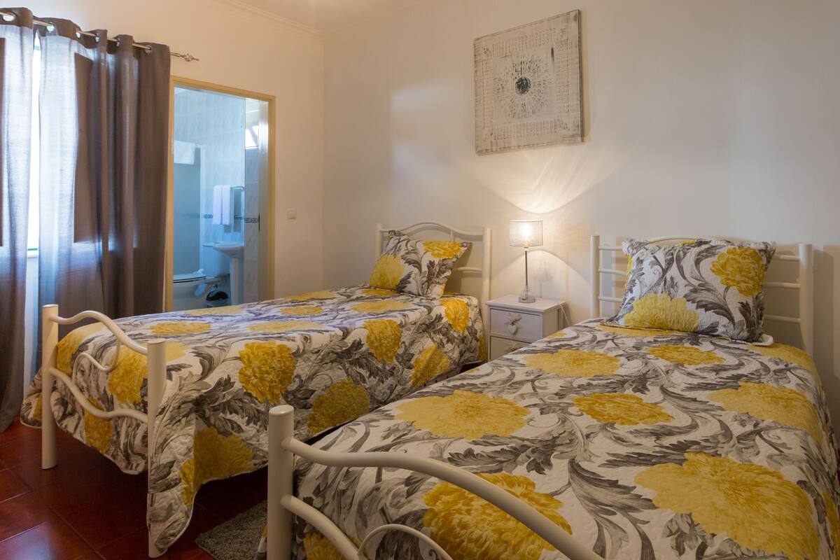 A bedroom with two beds, a bedside table with a lamp, and a bathroom visible through an open door.