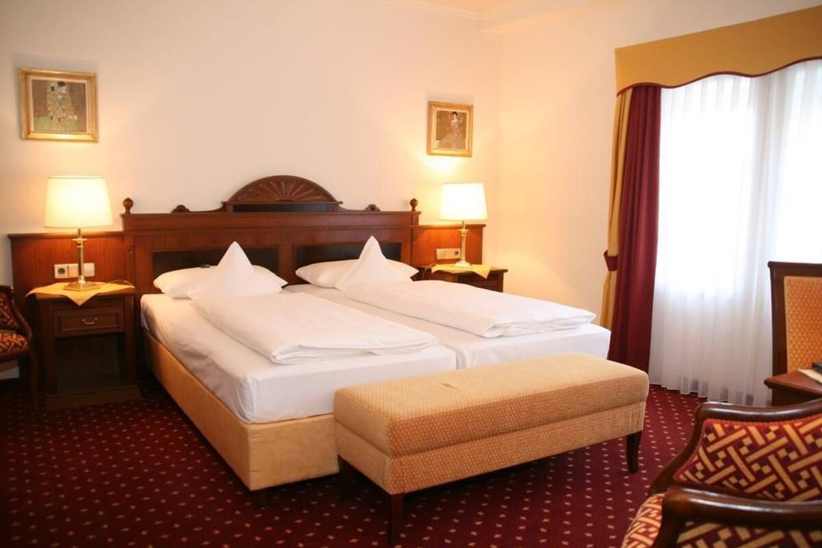 Habitación de hotel con cama doble, dos mesitas de noche con lámparas, un banco, una silla y un amplio ventanal con cortinas.