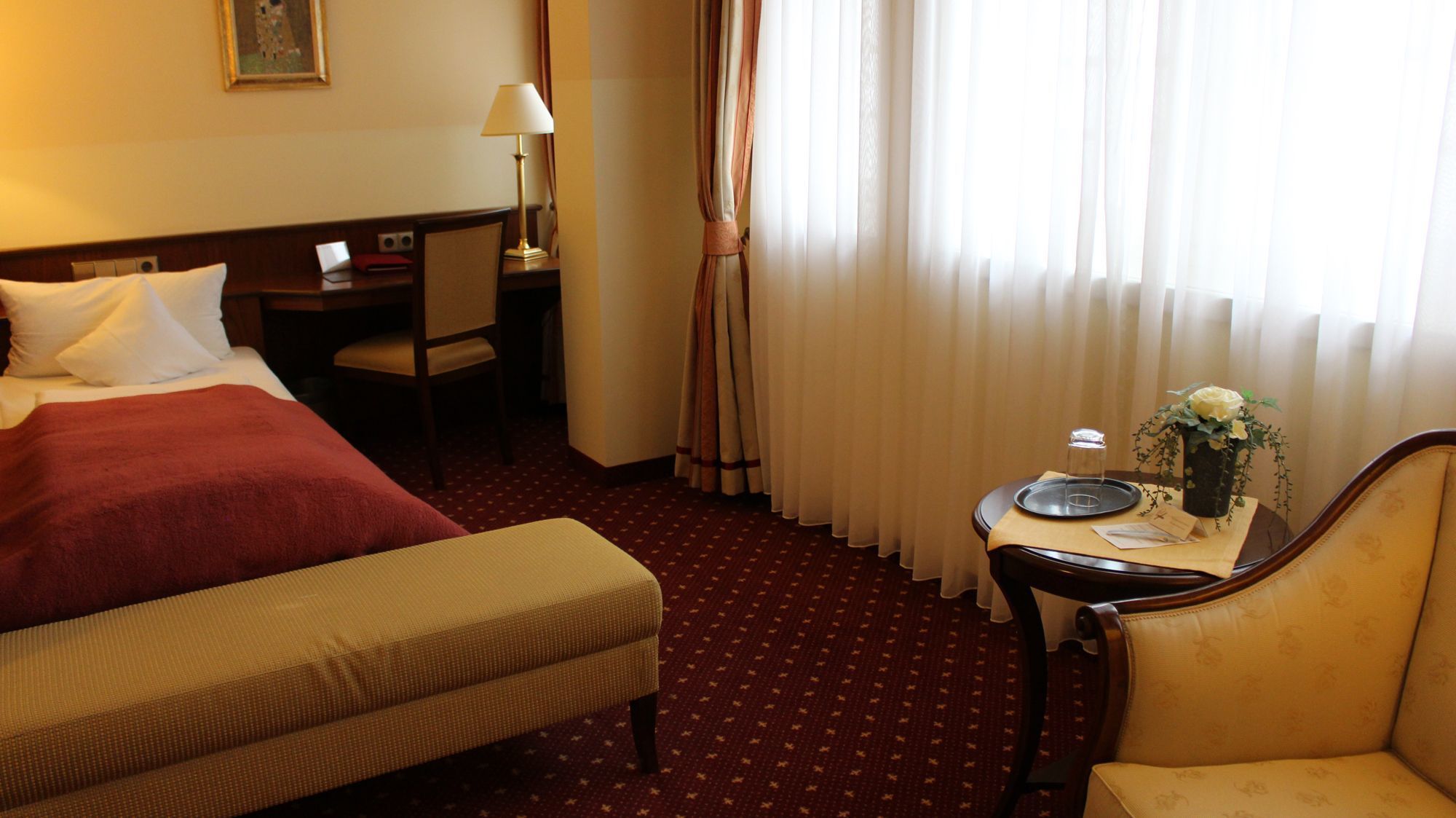 A hotel room with a bed, a desk, a chair, a small table with a vase and a tray, and a window with curtains.