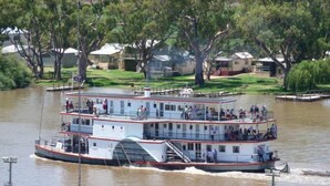 Un grand bateau à vapeur à plusieurs ponts, avec des passagers sur le pont supérieur, et un paysage fluvial paisible, avec des arbres et des bâtiments en arrière-plan.