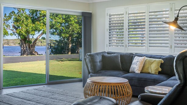 A living room with a grey sofa, a round wicker coffee table, and a view of a lake through large glass doors. - 2-18 Mangrove Street (EVANS HEAD)