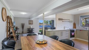 A modern kitchen with a large island, a dining area with a wooden table and chairs, and a view of the outdoors through a large window. - 2-18 Mangrove Street (EVANS HEAD)