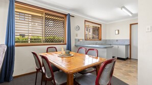 A dining area with a wooden table and chairs, a kitchen with a sink and cabinets, and a window with blinds. - 1-44 Woodburn Street - (EVANS HEAD)