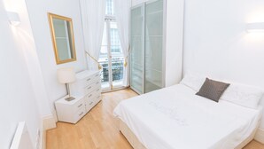 Quarto com cama, cômoda, abajur e vista para o exterior pela janela.