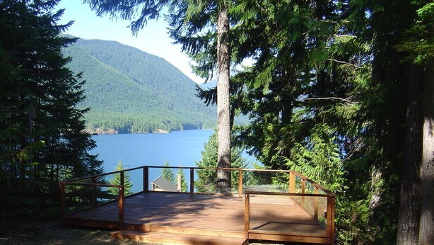 A wooden deck with a railing overlooking a lake, surrounded by dense forest and mountains.