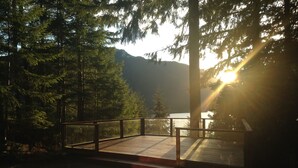 A wooden deck with a glass railing overlooking a lake, surrounded by trees and mountains.