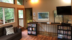 A cozy living room with a wooden ceiling, a large window overlooking trees, a bookshelf, and a flat-screen TV.