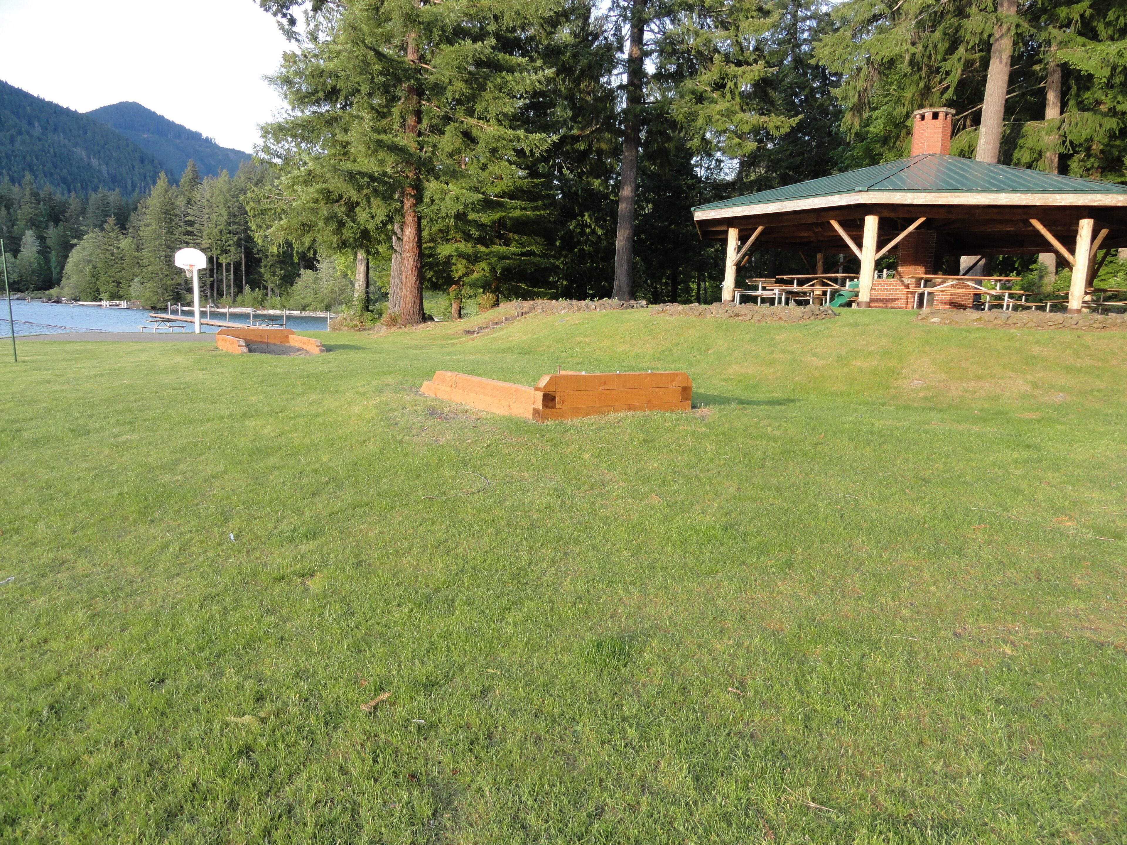 A grassy field with a wooden structure, a basketball hoop, and a body of water in the background.