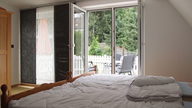 A bedroom with a bed, a wooden wardrobe, a sliding glass door leading to a balcony with a white fence and chairs. - Ferienwohnung Villa Am Golfplatz (Bad Harzburg)