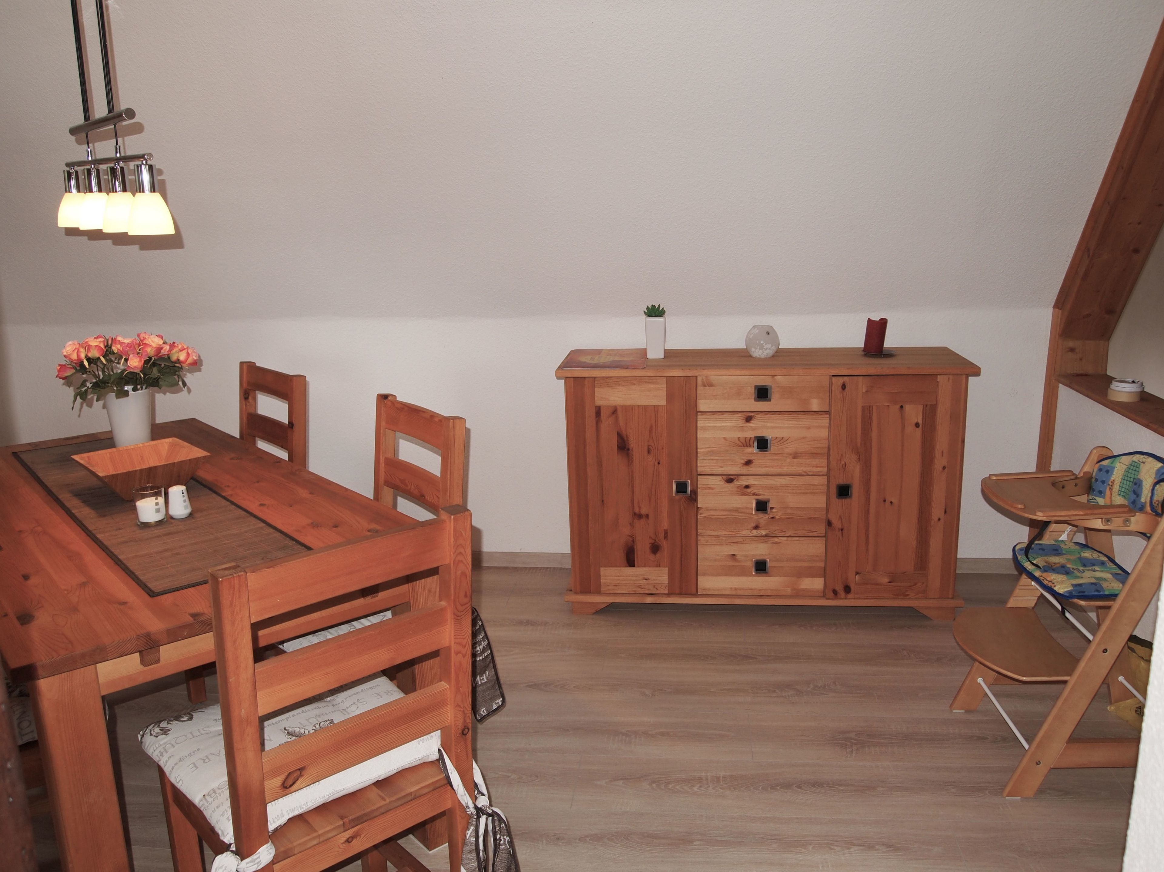 Un coin repas avec une table et des chaises en bois, un meuble avec des tiroirs, une petite plante et une chaise haute pour enfant.