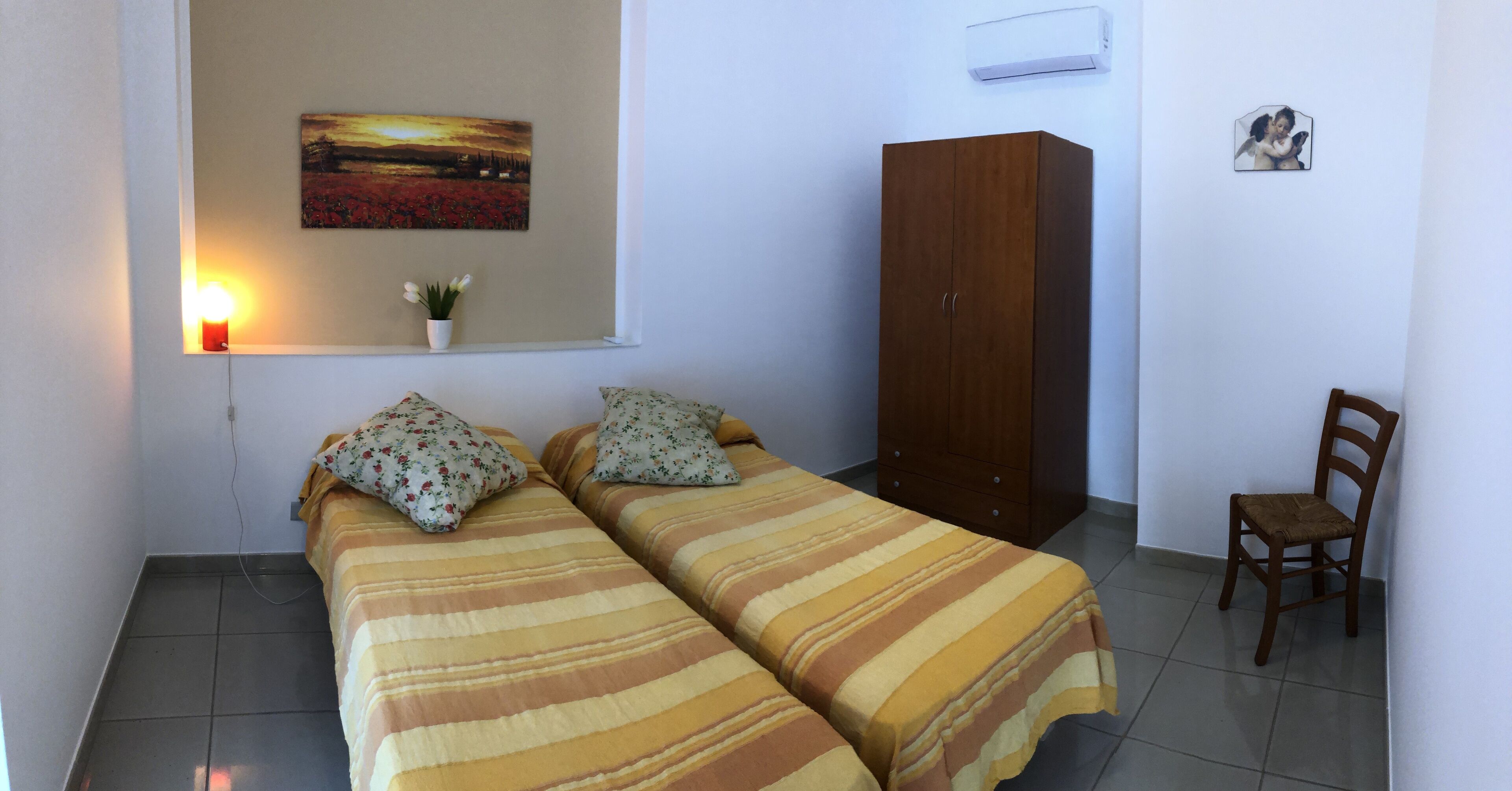 Quarto com duas camas, um guarda-roupa de madeira, uma cadeira, ar-condicionado fixado na parede, um quadro e uma mesinha com vela e flores.