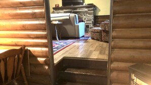 Interior de una cabaña de troncos con chimenea, un sofá, una alfombra estampada y una escalera de madera.