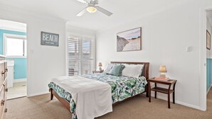 A bedroom with a bed, a nightstand, a ceiling fan, a window with blinds, a beach-themed sign, and a framed picture on the wall.
