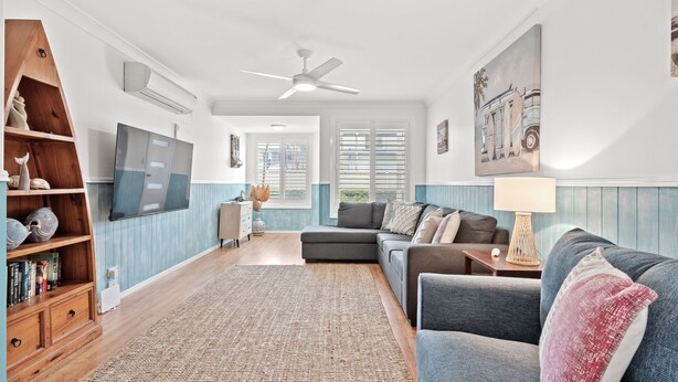 A living room with a sectional sofa, a TV mounted on the wall, a ceiling fan, and a large window with shutters.
