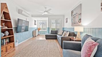 A living room with a sectional sofa, a TV mounted on the wall, a ceiling fan, and a large window with shutters.