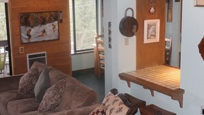 Een gezellige woonkamer met een bruine bank, gekleurde kussens, een houten wand met een schilderij en een klein houten plankje.