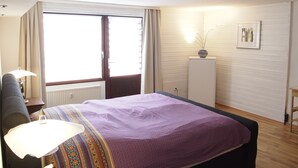 Een slaapkamer met een bed, nachtkastje, lamp, raam met gordijnen, radiator, kast en een ingelijste foto aan de muur.
