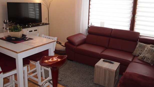 Een woonkamer met een televisie, een rode bank, een houten salontafel en een eethoek met een witte tafel en stoelen.