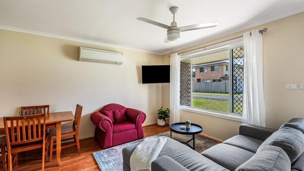 Ein Wohnzimmer mit Deckenventilator, einem an der Wand montierten Fernseher, einem Esstisch mit Stühlen, einem roten Sessel, einem grauen Sofa und einem Fenster mit Blick auf ein Haus.